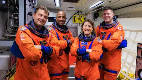 Frank Michaux/NASA Three men and a woman in orange astronaut uniforms smile with their arms folded as they face the camera.