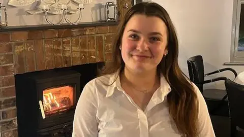 Chloe Claxton Willow is sitting wearing a white shirt in the pub she works in. She has long brown hair and a fireplace is visible behind her.