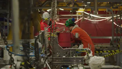 BBC workers in orange overalls operate heavy machinery